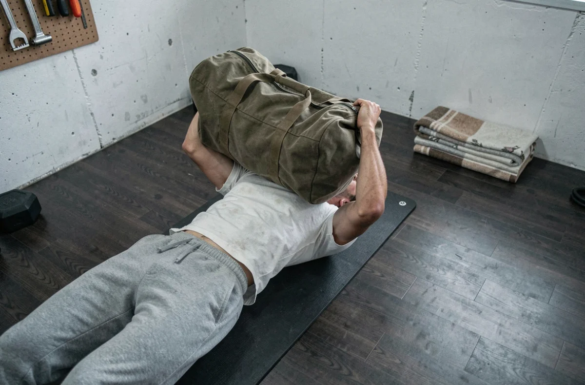 Homme réalisant un développé couché au sol avec un sac militaire lesté dans un garage aménagé