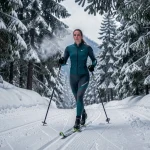 Femme pratiquant le ski de fond sur une piste damée dans une forêt de sapins enneigés en station nordique alpine